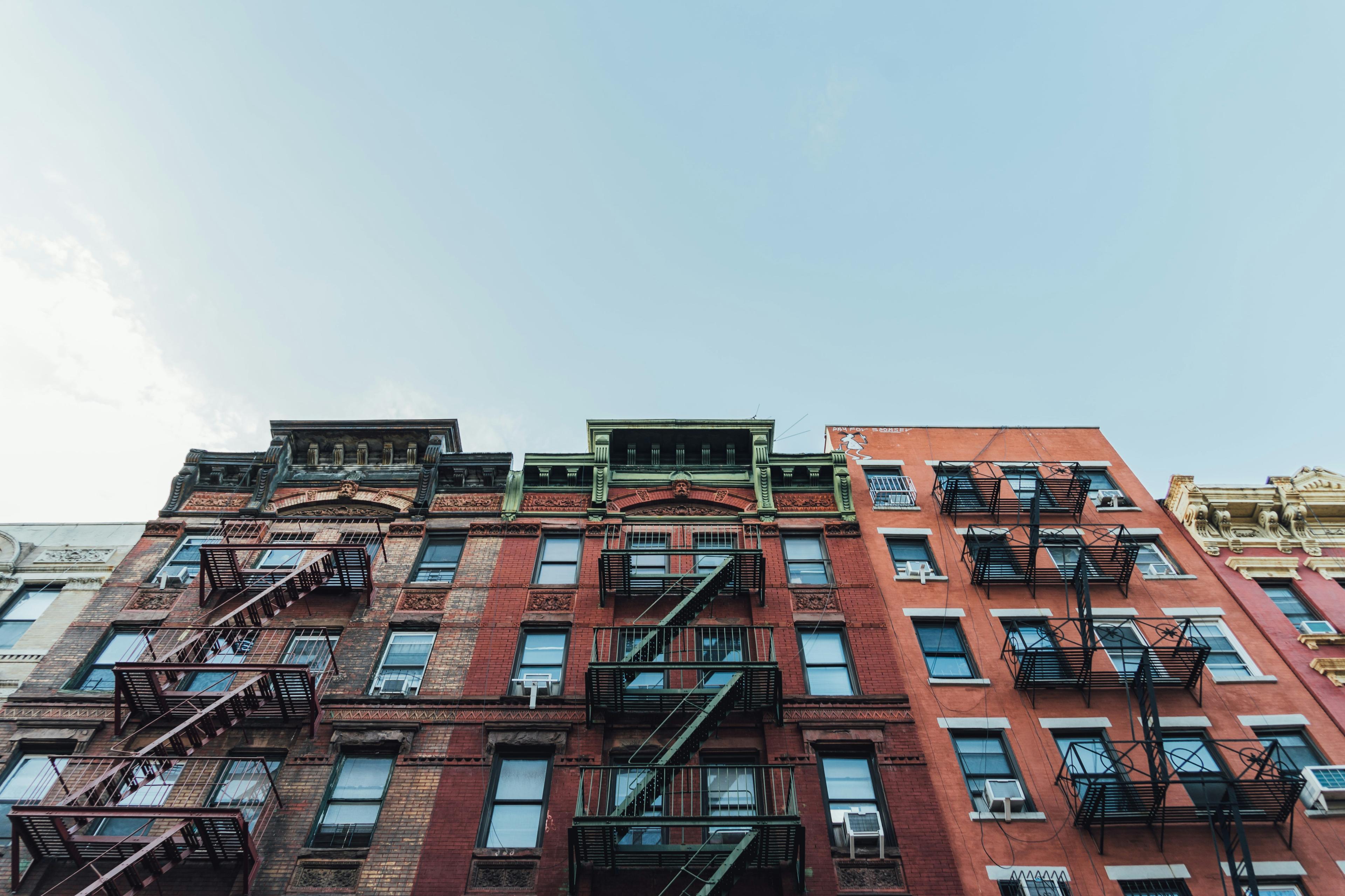 Sun-lit red-brick NYC tenement looking up from the street, fire escapes and decorative cornice visible