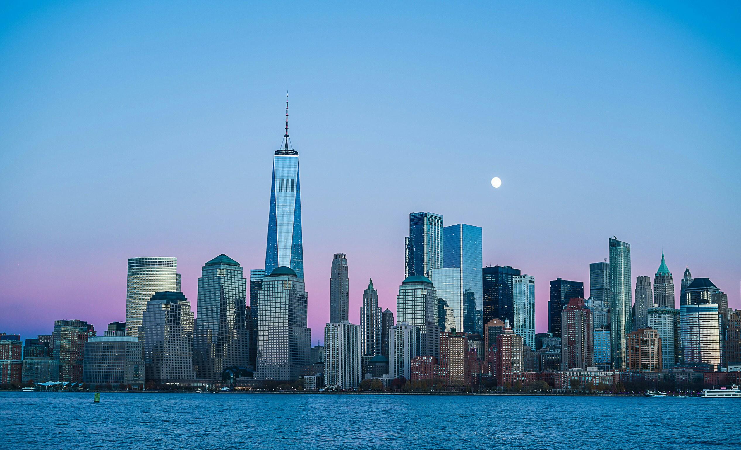 Downtown Manhattan skyline at dusk with One World Trade Center, a rising moon, and pink-purple sky reflected in the harbor