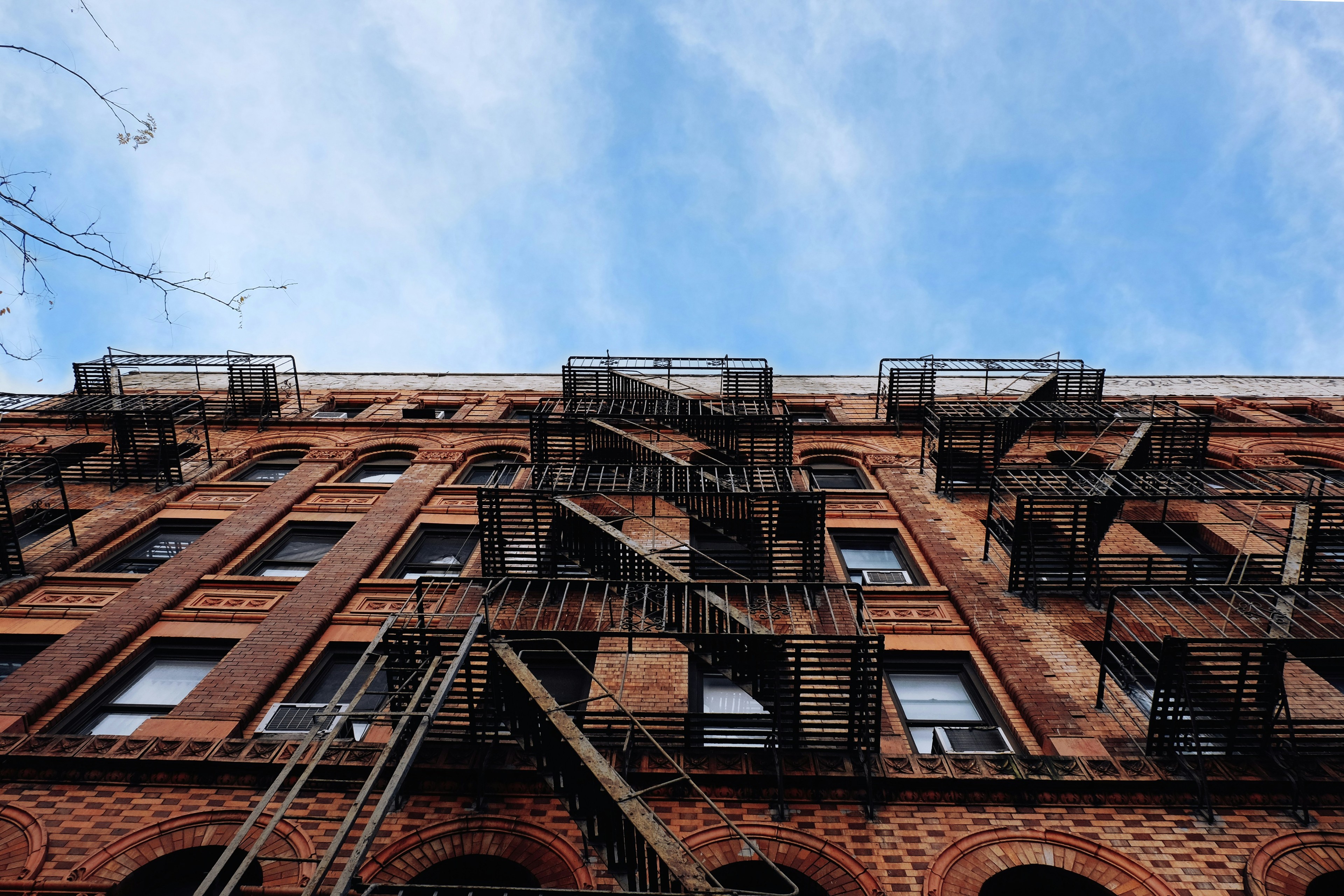 Looking up at a NYC red-brick tenement with crisscross fire escapes against a bright blue sky
