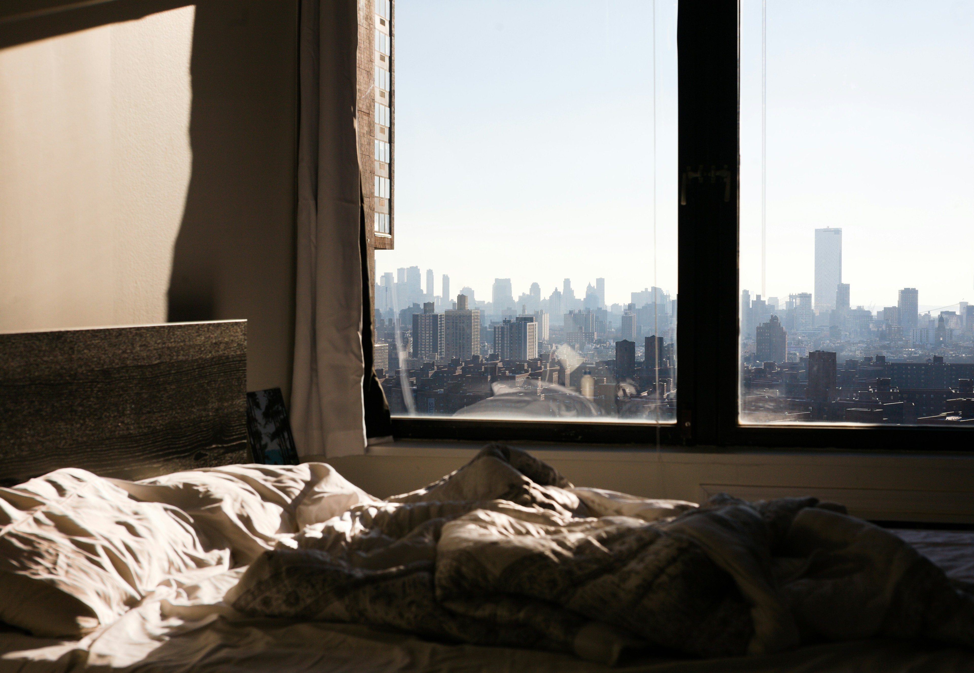 Sun-lit NYC bedroom with white rumpled sheets in the foreground and the Manhattan skyline visible through the open window